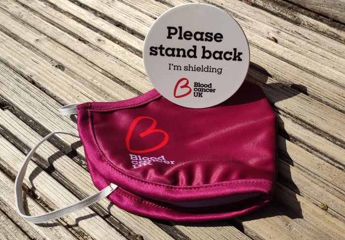 A Blood Cancer UK facemask placed on a table with a Blood Cancer UK please stand back I'm shielding badge, placed on top of it.