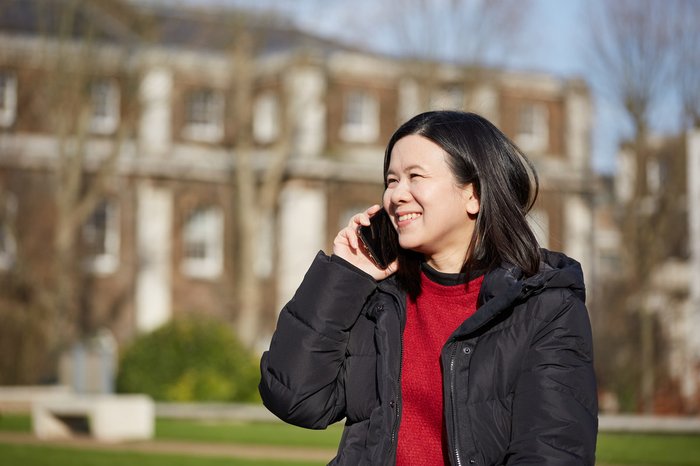 Jacqueline, in remission from DLBCL, smiles whilst holding a phone to her ear, standing outside in front of a house.