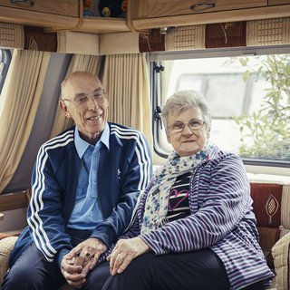 Trevor, who has chronic lymphocytic leukaemia (CLL), sits smiling, holding hands with his wife on the sofa at the back of a caravan.