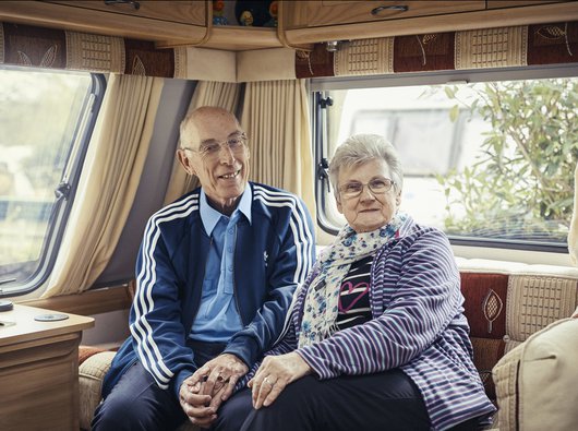 Trevor, who has chronic lymphocytic leukaemia (CLL), sits smiling, holding hands with his wife on the sofa at the back of a caravan.