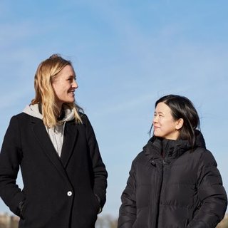 Jacqueline, in remission from DLBCL, stands smiling with her friend on a clear sunny day.