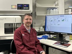 A man sat at a computer in a research lab wearing a red Blood Cancer UK lab coat.