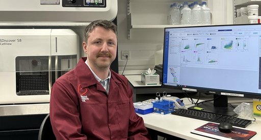 A man sat at a computer in a research lab wearing a red Blood Cancer UK lab coat.