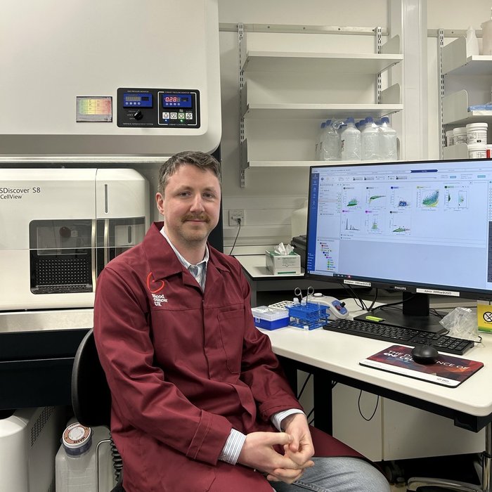 A man sat at a computer in a research lab wearing a red Blood Cancer UK lab coat.