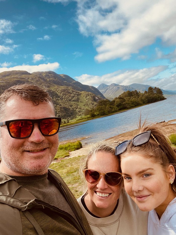Ally with his wife and daughter. They are standing together outside, with water and hills in the background.