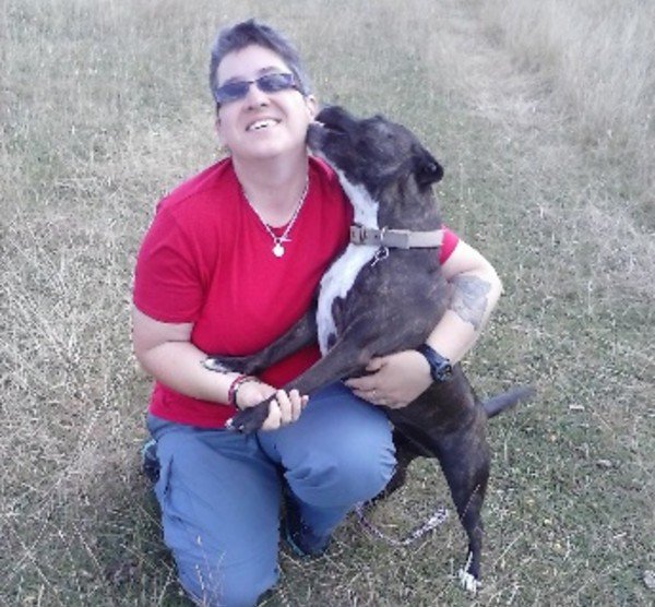 Angie Sharp who lives with CML, smiles whilst kneeling down, getting licked on her face by a dog.