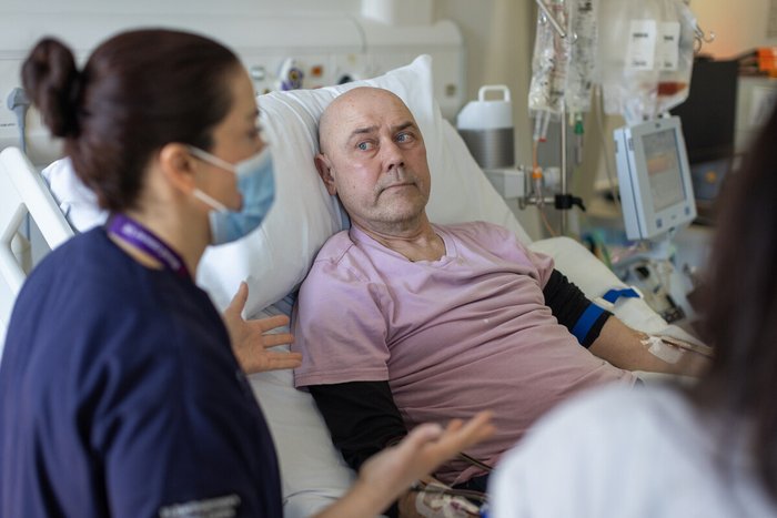 A nurse in navy scrubs, wearing a face mask, talking to a patient lying in a hospital bed.