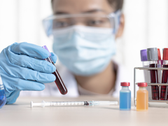 A researcher wearing a facemask inspects a vial of blood on a table with other vials.