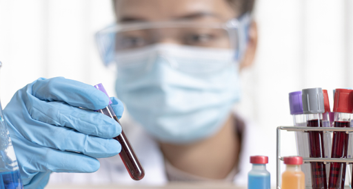 A researcher wearing a facemask inspects a vial of blood on a table with other vials.
