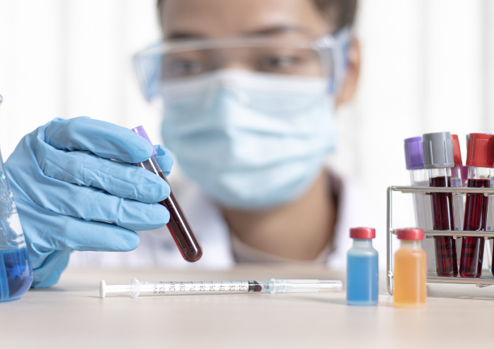 A researcher wearing a facemask inspects a vial of blood on a table with other vials.