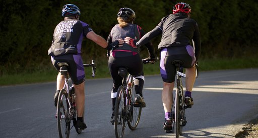 Three cyclists pedal up a hill, the cyclist on either side support the middle cyclist by patting them on the back.