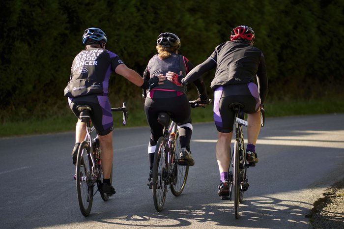 Three cyclists pedal up a hill, the cyclist on either side support the middle cyclist by patting them on the back.