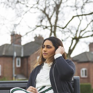 A young woman with blood cancer sits on a bench looking pensive, in front of a row of houses.