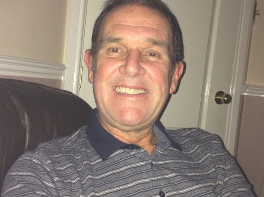 Brian, who lives with blood cancer, smiles to the camera whilst sitting at home on the sofa.