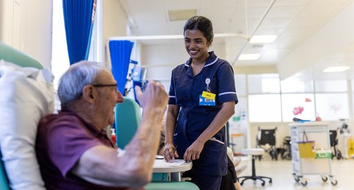 A female nurse and a male patient chat in the hospital. They are both smiling. The man is sitting in a chair, and the nurse is standing beside him.