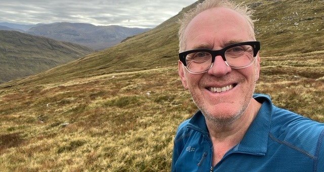 Colin taking a selfie on a grassy hillside with mountains behind him.