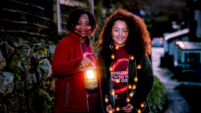 Two people smiling and holding lanterns under a moon-lit sky at a Walk of Light event.