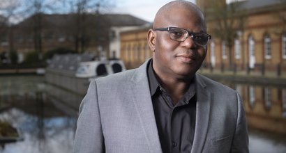 David, wearing a grey suit jacket and black shirt which is open at the neck. Behind David there is a canal boat and building.