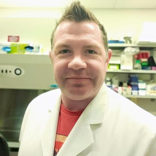 A headshot of a man with spikey hair wearing a white lab coat in the lab.