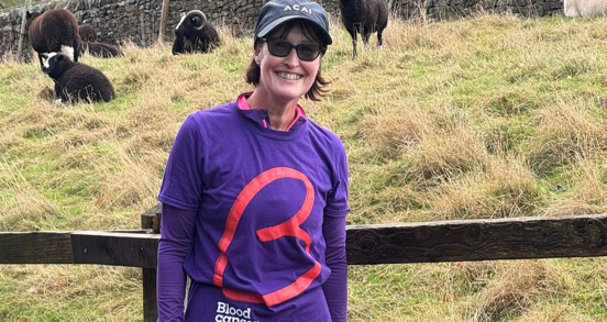 Denise wearing a purple Blood Cancer UK t-shirt, stood against a fence with sheep behind her