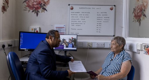A male doctor and a female patient sit opposite eachother in clinic. The doctor is handing the patient some written information.