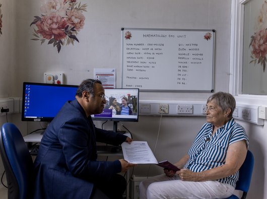 A male doctor and a female patient sit opposite eachother in clinic. The doctor is handing the patient some written information.