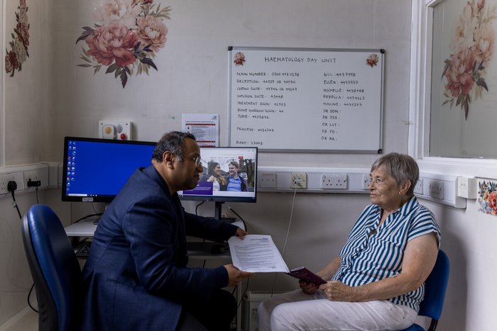 A male doctor and a female patient sit opposite eachother in clinic. The doctor is handing the patient some written information.