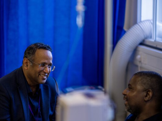 Doctor speaking to patient behind a curtain, there is an infusion pump in the forefront of the camera.