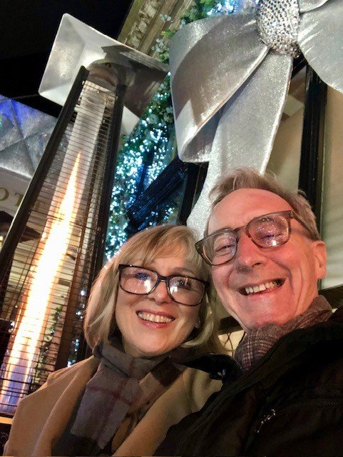 Doug and his wife Di, on a street at night. Buildings decorated with Christmas decorations are in the background.