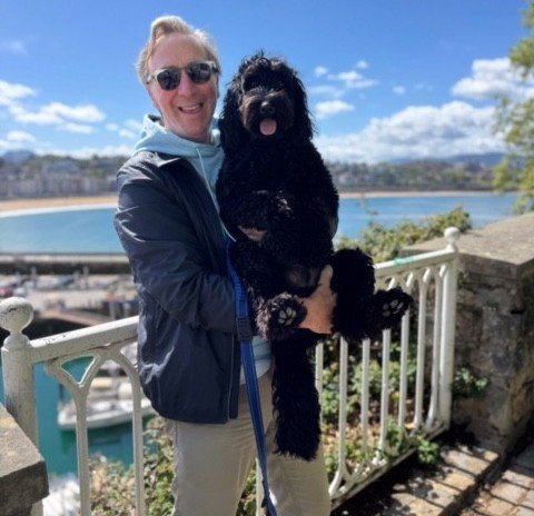 Doug, who had acute myeloid leukaemia, standing on a terrace in the sunshine with a lake or sea in the background. He's smiling, and holds a shaggy black dog in his arms.