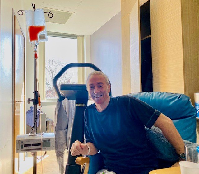 Doug in hospital on the day of his stem cell transplant. He's sitting in a chair in a hospital room. To his side, an IV bag on a stand contains pinkish red fluid: the new stem cells.