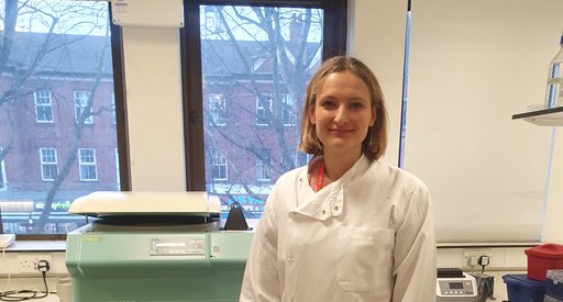 A women stood in the lab smiling wearing a white lab coat.