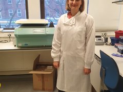 A women stood in the lab smiling wearing a white lab coat.