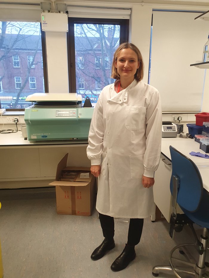 A women stood in the lab smiling wearing a white lab coat.