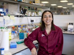 Dr Jasmeen Oberoi stood smiling in the lab wearing a Blood Cancer UK lab coat.