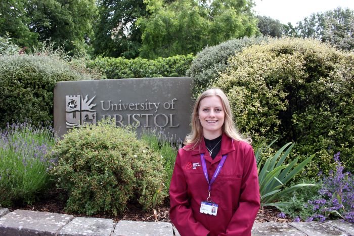 A lady stood outside by a University of Bristol sign wearing a red Blood Cancer UK lab coat.