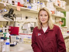 A lady with blonde hair stood in a science lab wearing a red Blood Cancer UK lab coat.