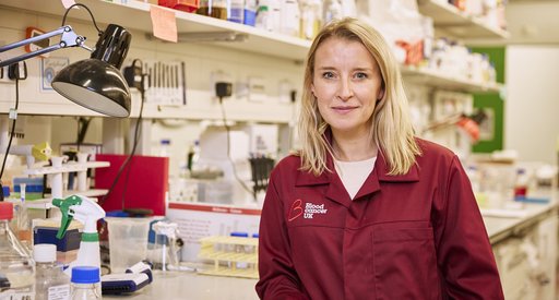 A lady with blonde hair stood in a science lab wearing a red Blood Cancer UK lab coat.