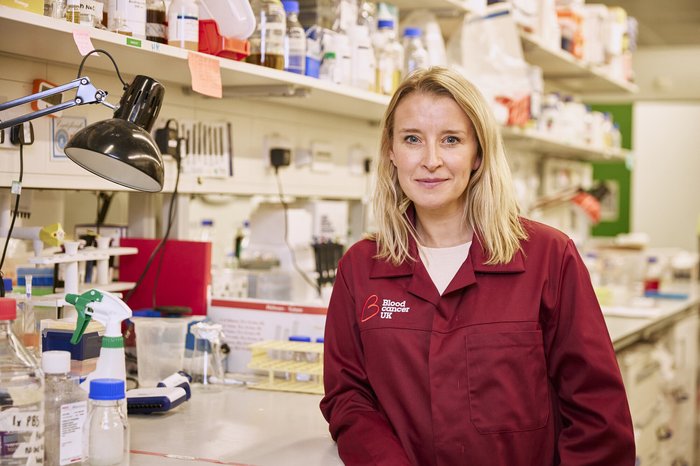 A lady with blonde hair stood in a science lab wearing a red Blood Cancer UK lab coat.