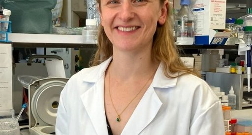 Image of a lady with light hair standing in a lab, smiling at the camera, wearing a white lab coat.