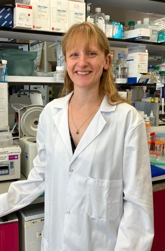 Image of a lady with light hair standing in a lab, smiling at the camera, wearing a white lab coat.