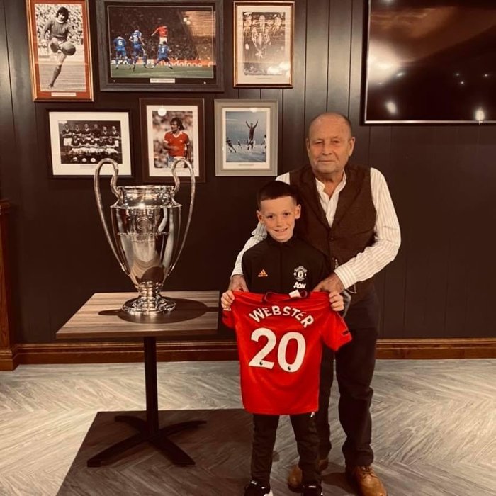 A young boy poses with an older man next to a football trophy, holding a jersey with WEBSTER 20 written on it.