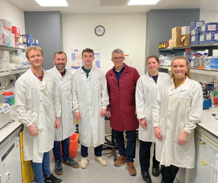 Professor George Vassiliou wearing a red Blood Cancer UK lab coat stood smiling in the lab with 5 members of his team who are in white lab coats