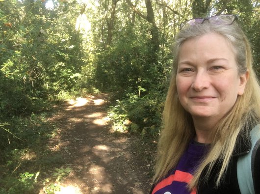 Gill, who has blood cancer and is a patient representative for Blood Cancer UK smiling stood in a woodland on a sunny day.