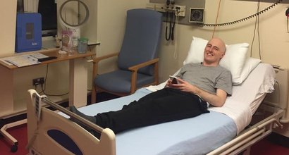 Graeme is lying on his back, on his hospital bed, with his head propped up on the pillow. He is looking at the camera and smiling.