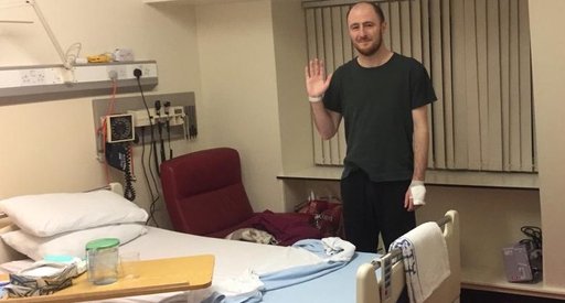 Photo of Graeme standing up behind his bed in his hospital room. He is smiling, and you can see a bandage around one hand, and a hospital wrist-band on the other hand.