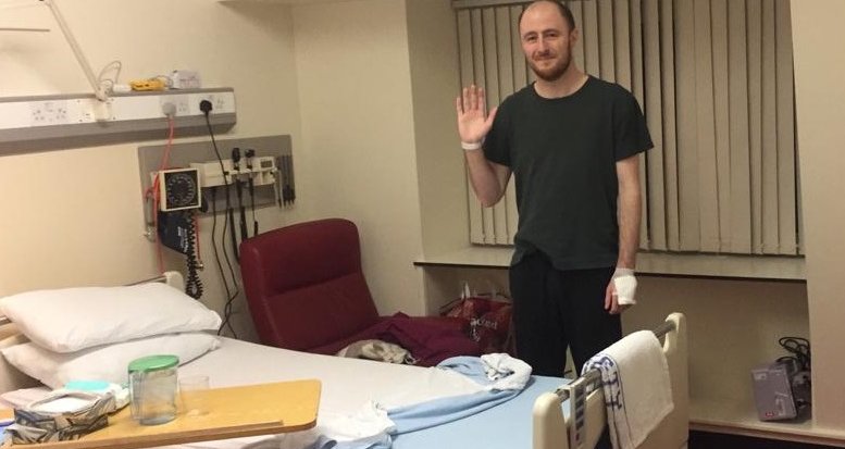 Photo of Graeme standing up behind his bed in his hospital room. He is smiling, and you can see a bandage around one hand, and a hospital wrist-band on the other hand.