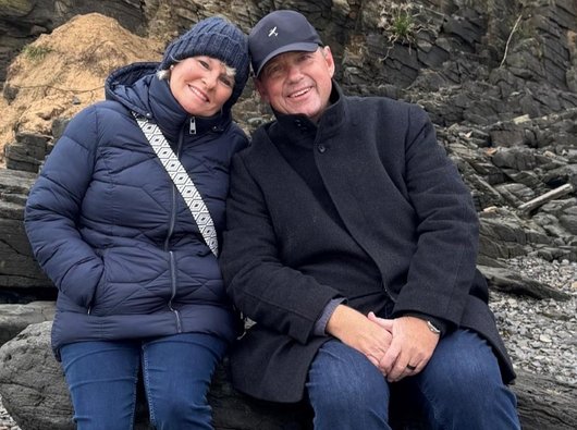 A photo of Grahame and his wife Nadine on a walk by the beach in winter. They are wrapped up warmly and relaxing against a large rock on the sea shore