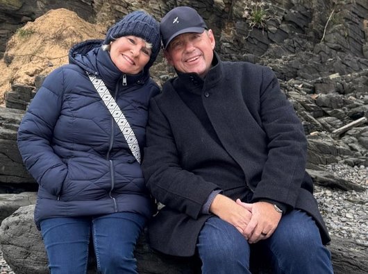 A photo of Grahame and his wife Nadine on a walk by the beach in winter. They are wrapped up warmly and relaxing against a large rock on the sea shore