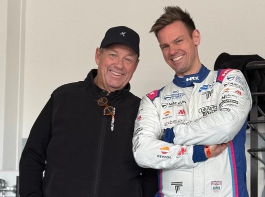 A photo of Grahame with his son Tom. Tom is wearing a racing driver's jumpsuit and they are standing by the bonnet of a racing car